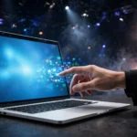 Sleek aluminium laptop on a desk with a hand reaching to touch its glowing screen under dark stage lighting